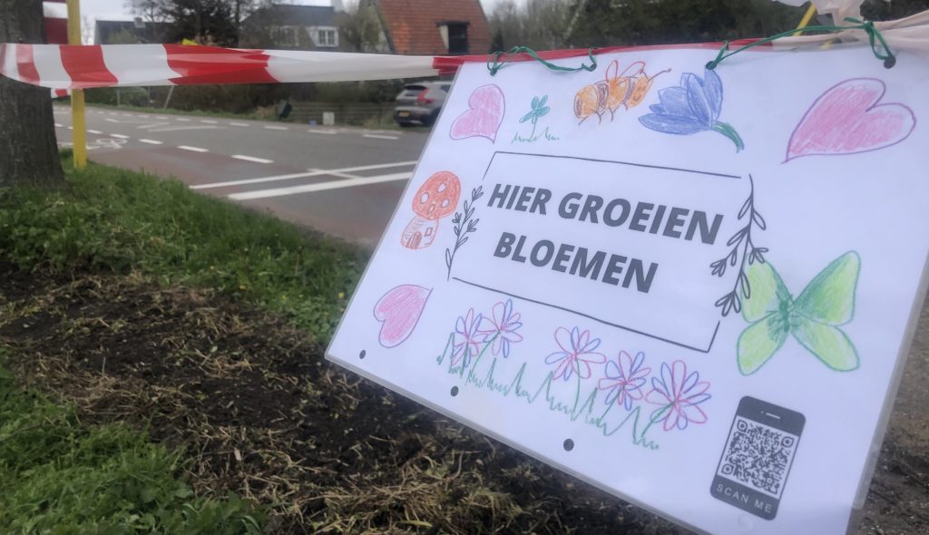 Op de achtergrond een landelijke weg met voorop een horizontaal gespannen gevarenlint met hieraan vastgemaakt een geplastificeerd document met tekeningen van bloemen en bijen en de tekst "hier groeien bloemen"